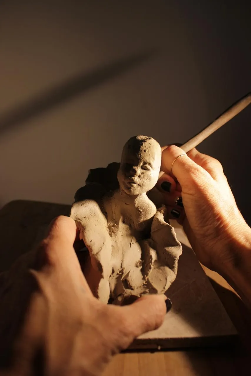 Hands shaping a small clay figure with a modelling tool, in warm light