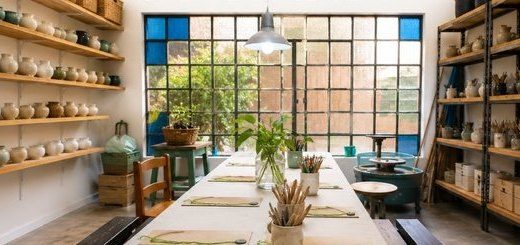 El espacio del taller: mesa larga de trabajo, estantes con piezas de cerámica y un gran ventanal