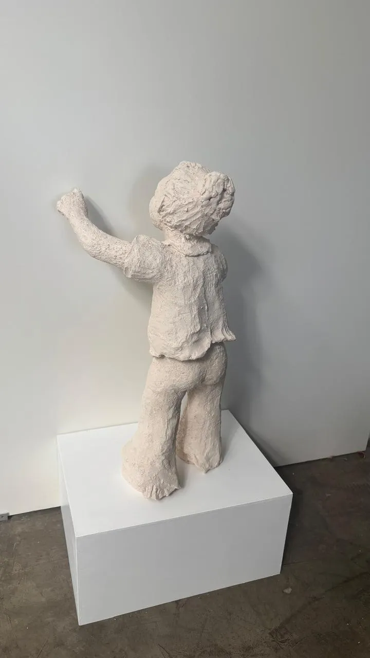 Tiempo circular: escultura de una niña de pie sobre un pedestal blanco, de espaldas, que escribe sobre la pared con el dedo