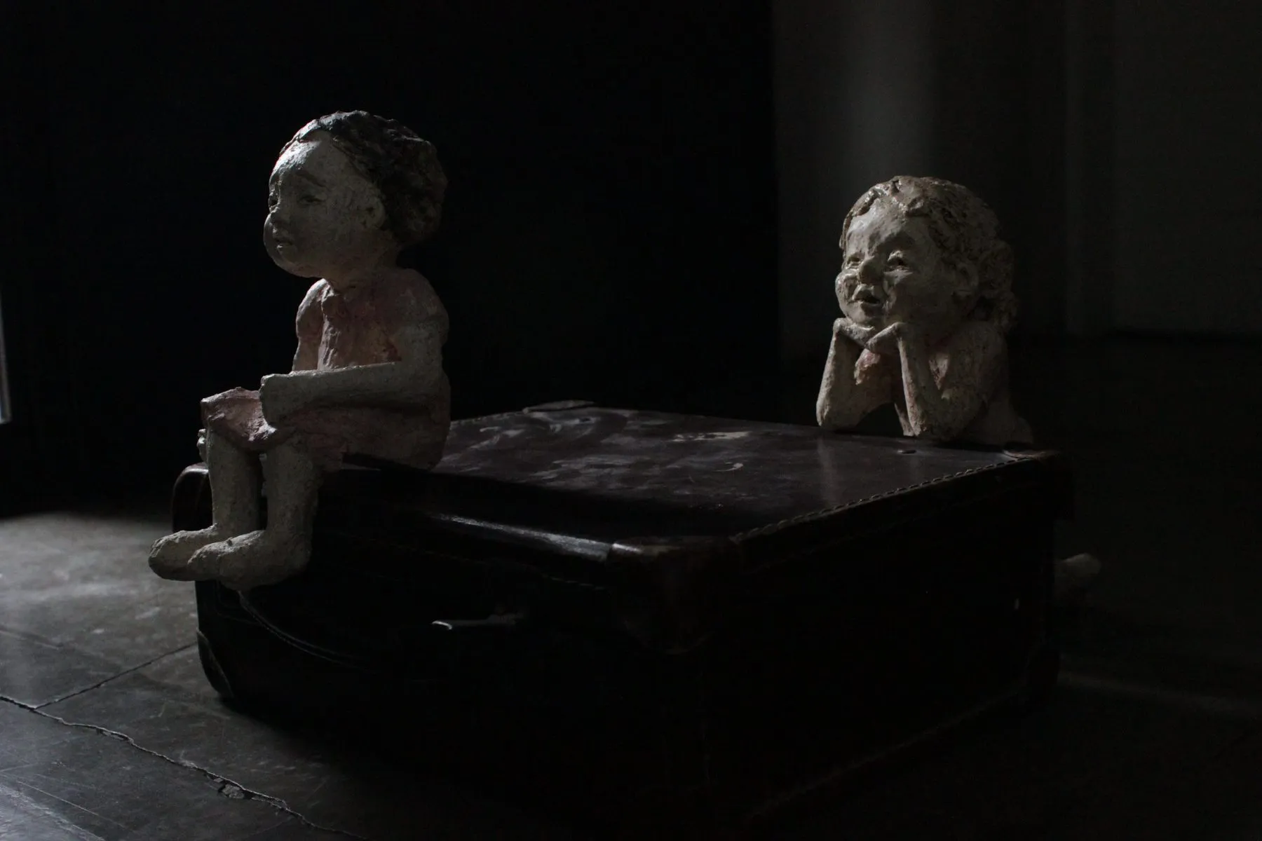 Trasmigrare: two ceramic children on an old suitcase, in semi-darkness