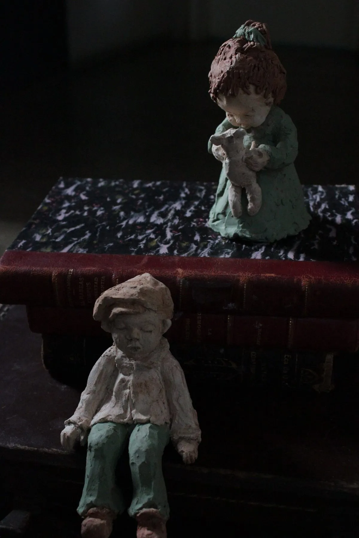 Two figures of children, a girl holding a doll and a seated boy, on books and a suitcase